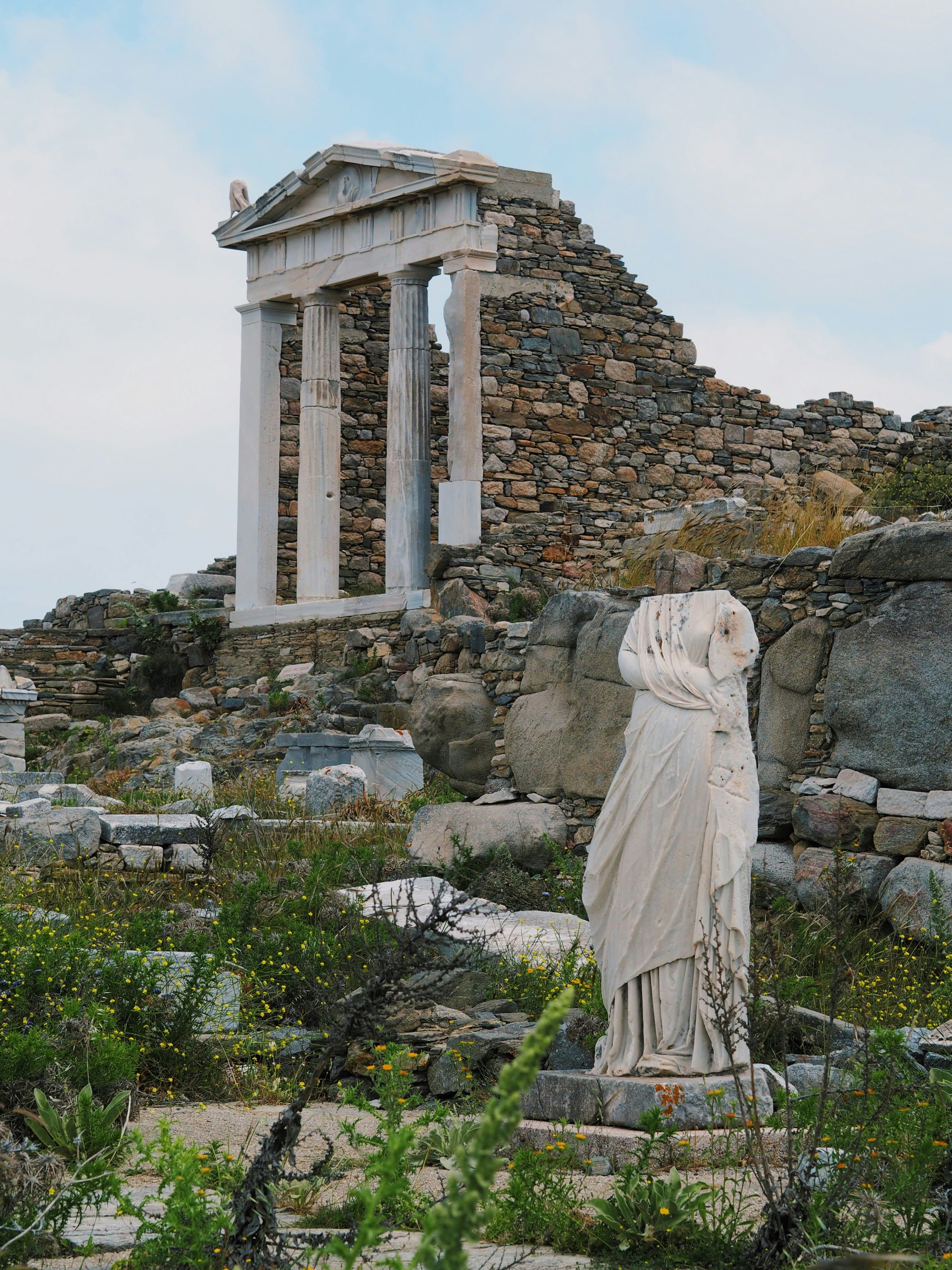 Ancient ruins of Delos Island featuring a headless marble statue and partially restored Hellenistic temple with stone walls in the background.