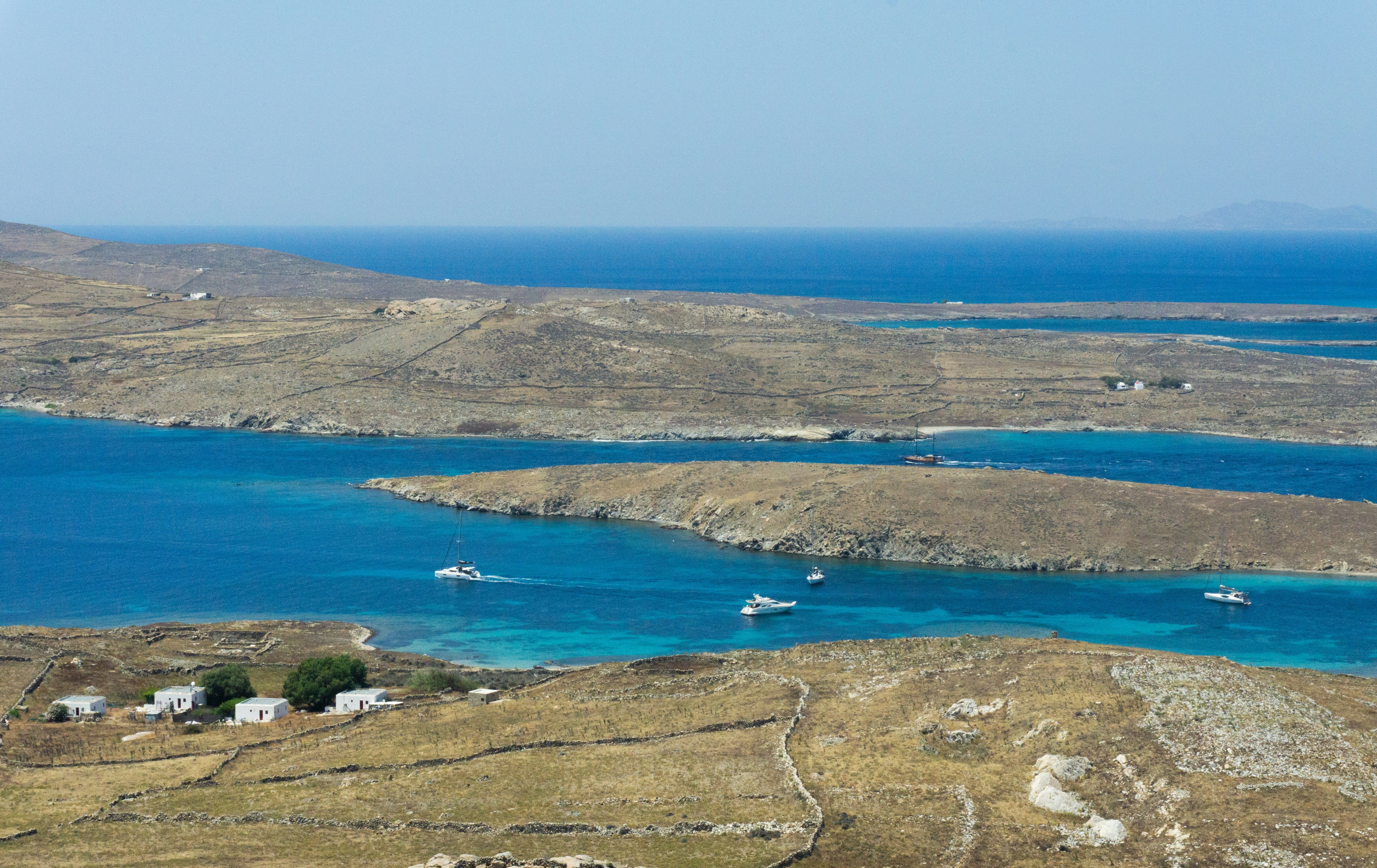 Aerial view of Delos Island’s coastline with turquoise waters, anchored sailing yachts, and dry, rugged Cycladic landscape.