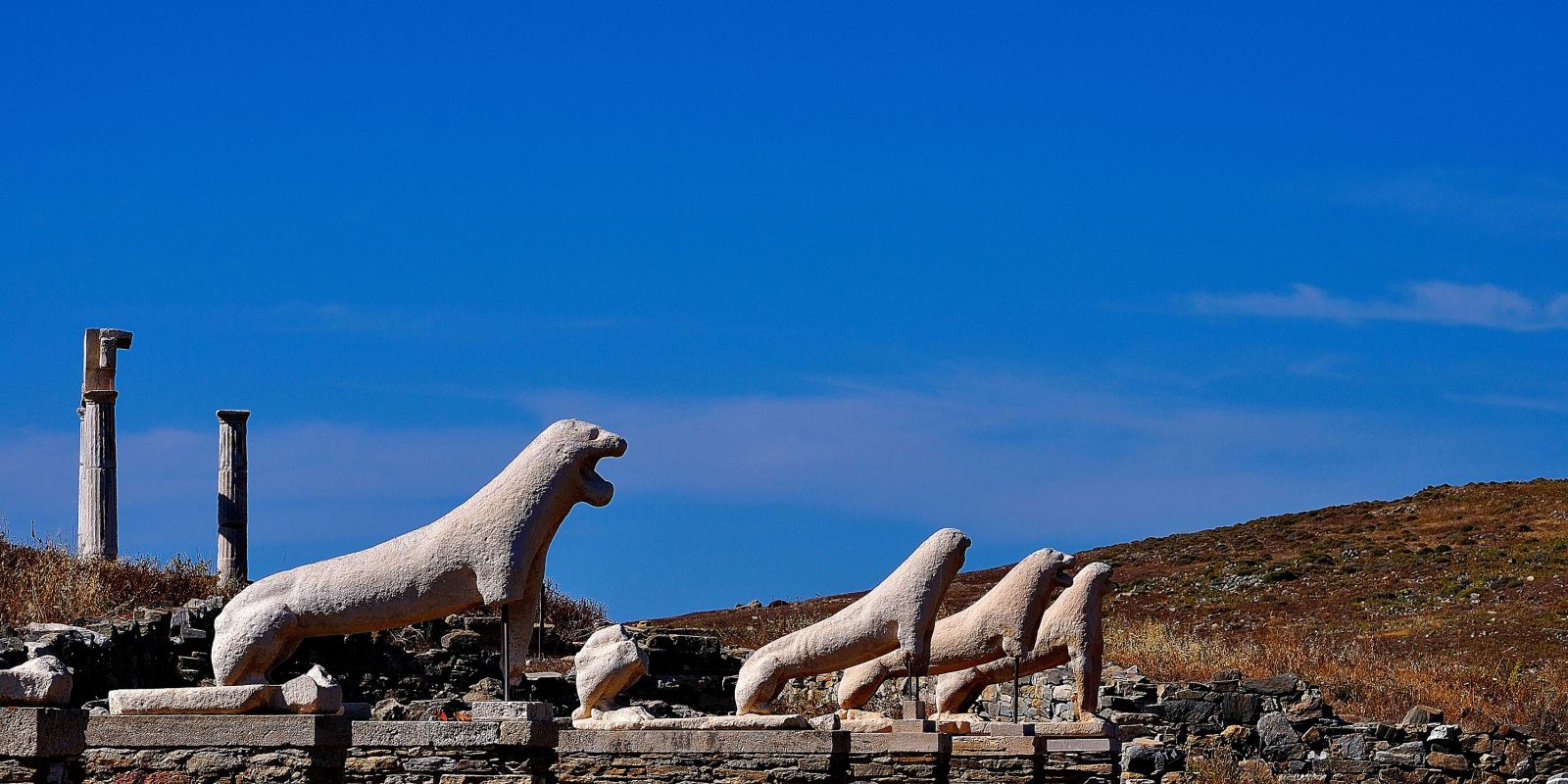 Ancient marble lion statues from the Terrace of the Lions stand in a row under a bright blue sky on Delos Island, Greece. Surrounded by weathered ruins and dry, rocky terrain, these iconic sculptures highlight the island's rich archaeological and mythological heritage.