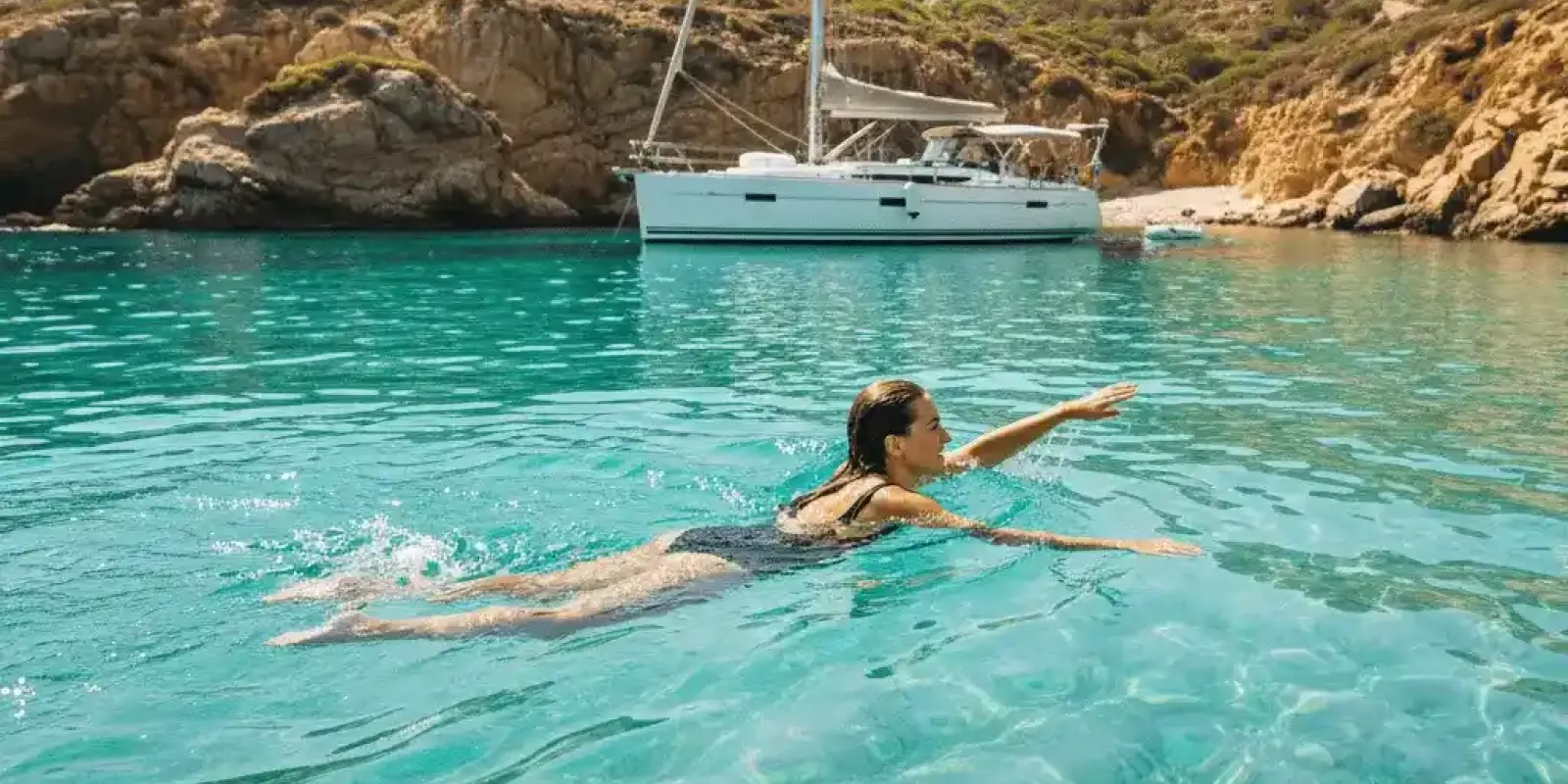 A person swimming in crystal clear turquoise waters off a beautiful, secluded cove, with a yacht visible in the background.