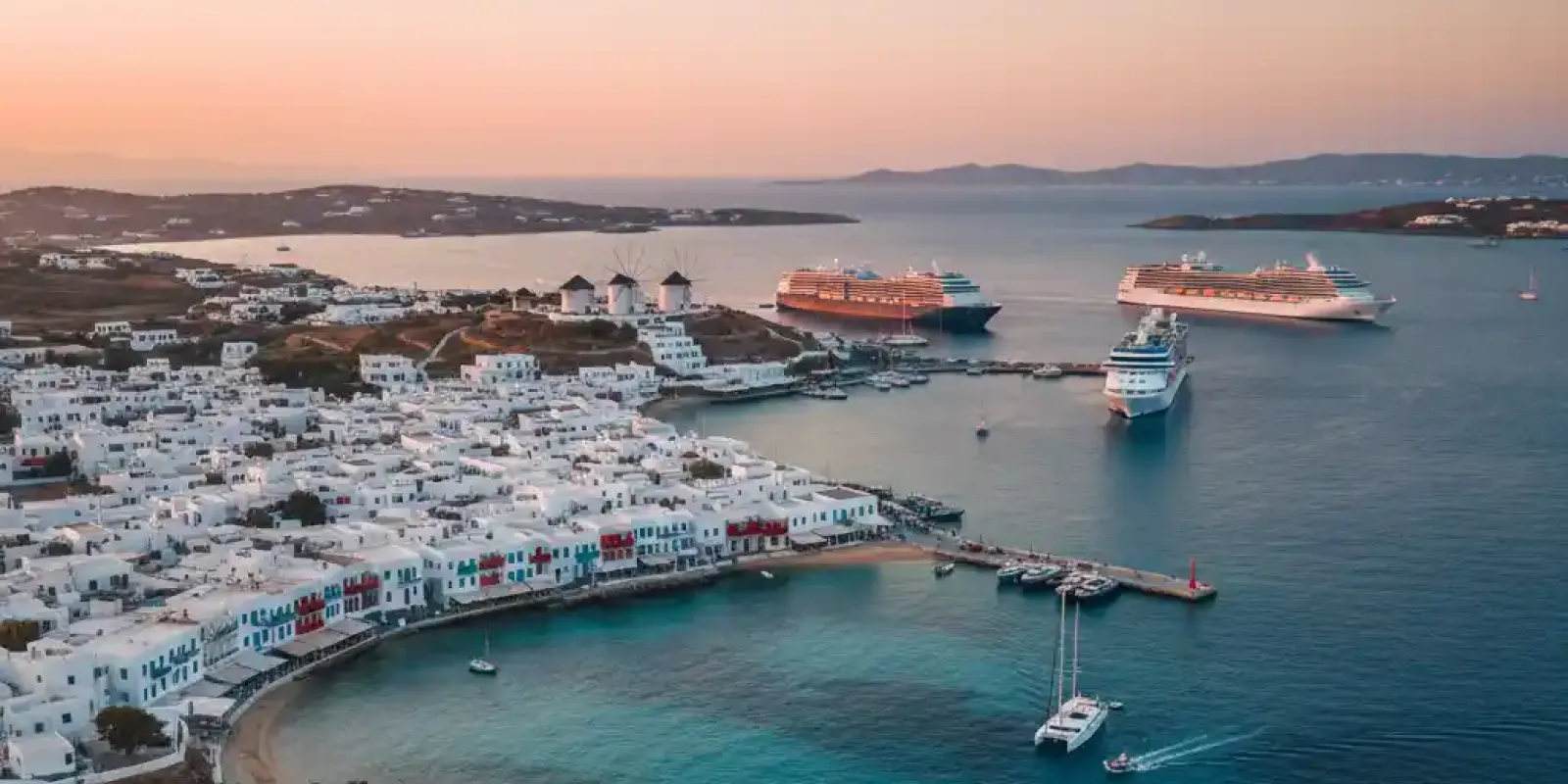 An aerial shot of Mykonos Town, showcasing its white buildings, coastal port, and several large cruise ships anchored in the bay during sunset.