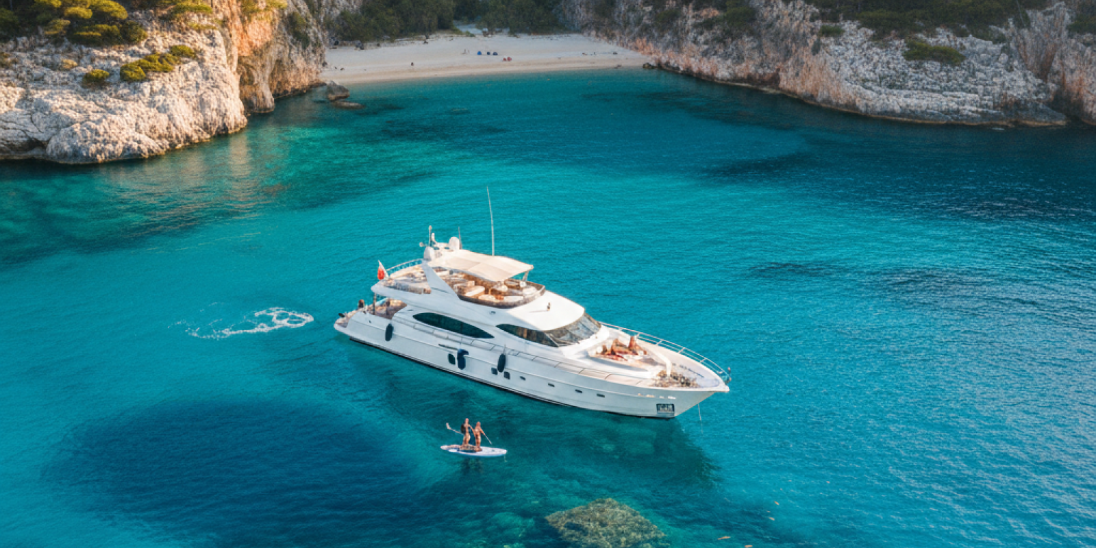 An aerial view of a luxury yacht anchored in a secluded, turquoise-water bay with pine-covered cliffs and a small beach, with a person paddleboarding near underwater ruins.